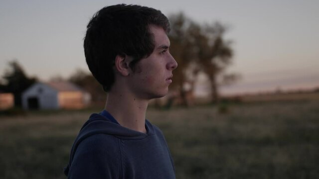 Side medium shot of a young, teenage boy watching the sun set in a Kansas field. The teen looks serious and has dark hair.
