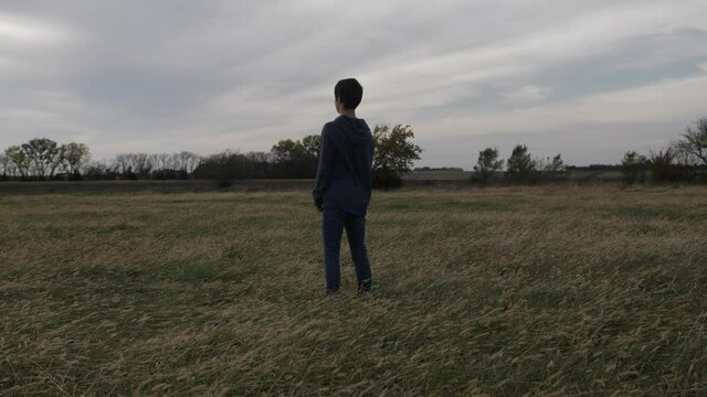 A teen boy stands in a Kansas field by himself watching the sun setting. The teenager is wearing jeans and a hoodie shirt and has dark hair.
