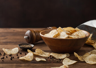 Crunchy potato crisps chips snack with black pepper in wooden bowl on dark background with mill and...