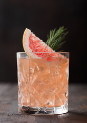 Red grapefruit summer juice cocktail in glass with rosemary and fruit slice on black background.