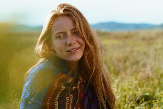 a girl with loose red hair in an ethno cape sits on a meadow against the backdrop of hills. close-up. High quality photo
