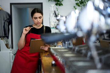 Serious female waitress in apron cafe uniform reading information from report checking accounting...