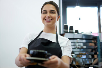 Portrait of cheerful caucasian woman barista in apron holding cup with hot aroma cappuccino at cafeteria, smiling 20s female waitress offering coffee beverage hospitably owning small business