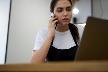 Serious female manager of coffee shop talking on mobile phone making online order on web page via...