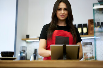 Half length portrait of skilled female cashier standing near digital tablet with application for...