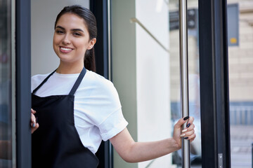 Half length portrait of smiling brunette waitress of coffee shop standing at entrance meeting visitors in morning, hospitable woman owner of cafeteria enjoying work and small business entrepreneurship