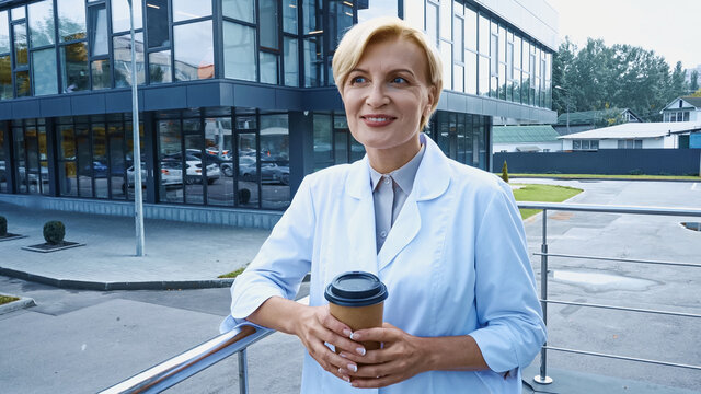 Smiling Doctor In White Coat Holding Coffee To Go Outdoors