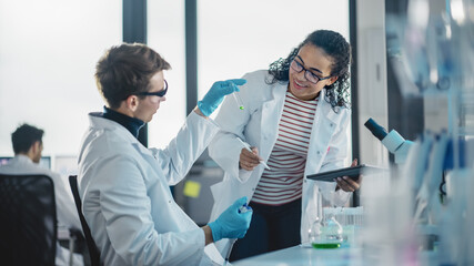 Modern Medical Research Laboratory: Portrait of Two Young Scientists Mixing Liquid Chemicals, Checking Result. Digital Tablet, Talking. Diverse Team of Multi-Ethnic Specialists work in Advanced Lab