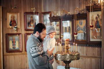 A Russian man with a beard and a daughter is standing in an Orthodox Church, lighting a candle and praying in front of the icon.