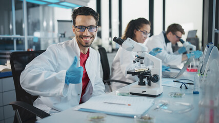 Medical Science Laboratory: Handsome Scientist is Using Microscope for Sample Analysis, Looking at Camera, Smiling Cheerfully, Shows Thumbs Up. Young Biotechnology Specialist, Using Advanced Equipment