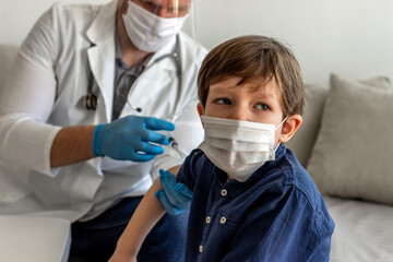 Selective focus of adorable little boy looking away, while male doctor holding a syringe and vaccinating him. A boy in a doctors office, looks concerned as he gets injected. Doctor vaccinating  boy.