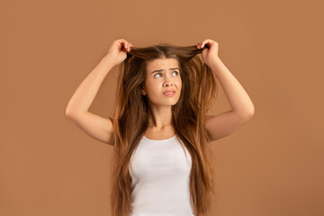 What a disaster. Upset young lady looking at her tangled long straight hair on brown studio...
