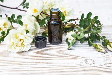 Black vial with essential rose oil and twig with white wild roses flowers on white paint wooden background. Space for text