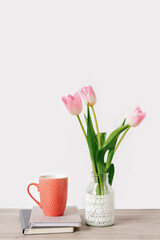A bouquet of three pink tulips stands in a glass vase with lace. Next to it are notebooks or notepads with a ceramic mug of tea.  Greeting card for Mother's Day. The concept of spring