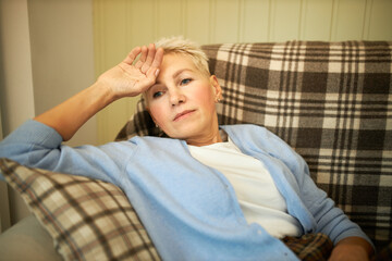 Frustrated sad female pensioner with short hair lying on sofa at home alone, touching forehead, having high temperature, suffering from cold or flu, recovering at home alone. People and health care