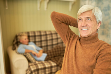 Obraz premium Handsome senior retired man wearing brown turtleneck sweater relaxing at home looking up with thoughtful dreamy smile, his wife having rest on couch in background. Quarantine and social distancing