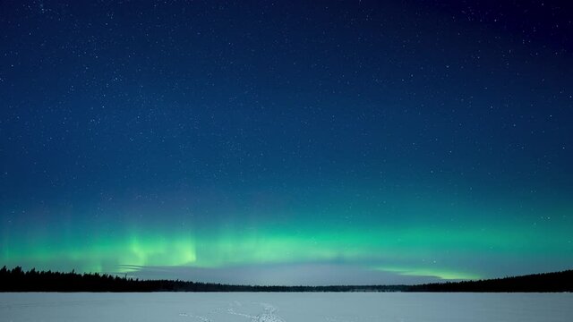 Time lapse of very active Aurora in a starry night sky with over a frozen snow covered lake and with a forest treeline in the distance.
