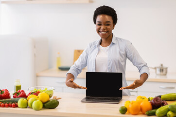 African Woman Demonstrating Laptop Screen Advertising Cooking Website In Kitchen