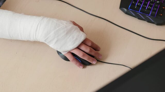 Broken arm in bandage clicks PC mouse while sitting at home close up top view. Hand of a teenager in a cast works with a computer