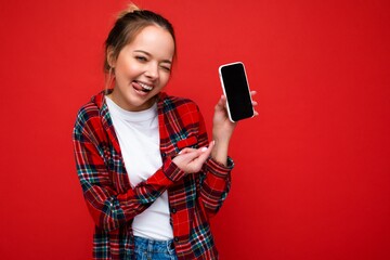 Photo of winking beautiful cute positive young blonde woman standing isolated over red wall wearing casual red shirt and white t-shirt showing mobile phone with empty screen for mockup looking at