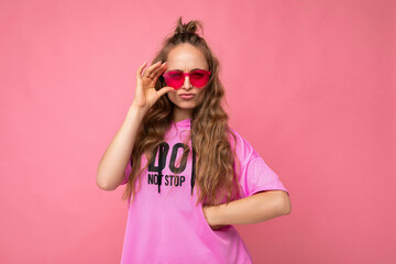 Photo shot of pretty sad dissatisfied young blonde curly woman isolated over pink background wall wearing casual pink t-shirt and stylish colourful sunglasses looking at camera and having questions