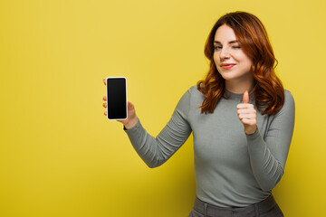 Naklejka premium cheerful woman with curly hair holding smartphone with blank screen and showing thumb up on yellow