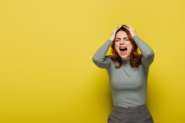 angry woman with wavy hair screaming and touching head on yellow