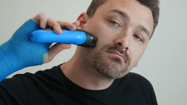 Man with hand wrapped in fiberglass plaster cast shaves his cheek with an electric safety razor. Male hand in blue waterproof bandage smoothly shaves stubble with a razor on a white background