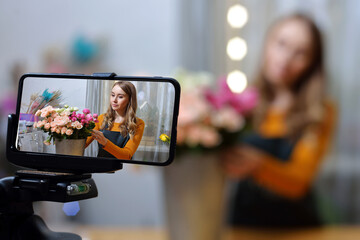 Small flower shop. A woman writes a video blog or online course. Flower school concept. Florist woman creates flower arrangement in a wicker basket.