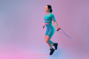 Fototapeta na wymiar Young fit and sportive woman jumps rope on gradient background. Fit sportswoman posing, looks confident. Perfect body ready for summertime. Beauty, resort, sport concept. Flyer