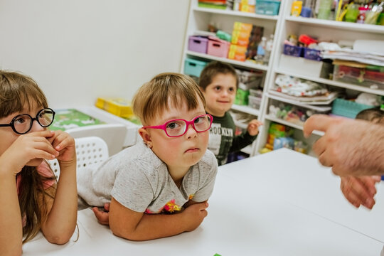 Young cute girl with down syndrome in grey shirt and pink glasses sitting at white desk with other kids and studying. 