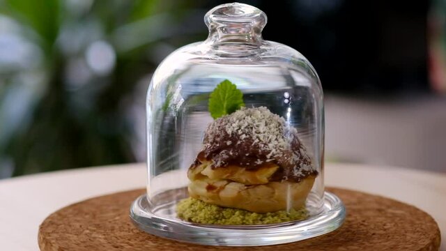 Inside of glass domed serving plate one portion of profiterole garnished with chocolate and coconut flakes, served on powdered pistachio nuts, close up spinning view. Restaurant dessert dish concept..