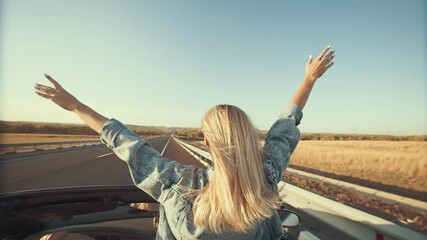 Woman with hands up outstretched stands in front seat of cabriolet, view from back. Blonde rides standing in convertible with her arms outstretched, enjoying wind and freedom of travel