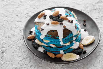 Vegan blue pancakes with sour cream and banana slices on a plate sprinkled with almonds, raisins on a gray textured background. Place to copy text