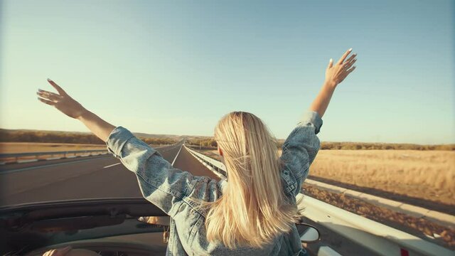 Woman with hands up outstretched stands in front seat of cabriolet, view from back. Blonde rides standing in convertible with her arms outstretched, enjoying wind and freedom of travel