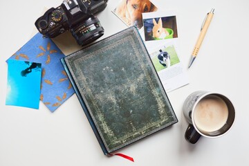 A beautiful notebook surrounded by a classical camera, some photos, a postcard, a pen and a coffee.