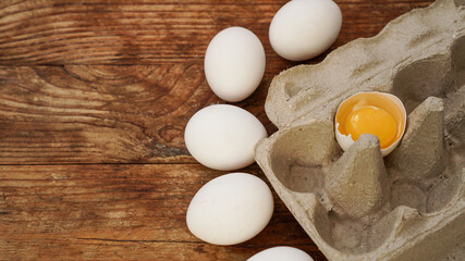 White eggs carton and cracked egg half with yolk top view on wooden background. Easter and healthy food breakfast cooking concept