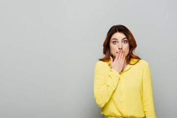 Thrilled woman in yellow blouse covering mouth with hand on grey