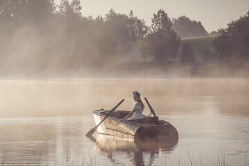 Beautiful girl in a light-blue dress rowing a boat on a misty lake during a sunrise.