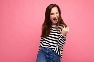 Portrait of young european emotional positive happy beautiful brunette woman with sincere emotions wearing casual striped pullover isolated on pink background with empty space and celebrating winning