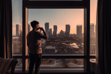 Young asian man standing with drinking coffee in a hotel. Sunrise over modern buildings in Songdo central park at Incheon - obrazy, fototapety, plakaty