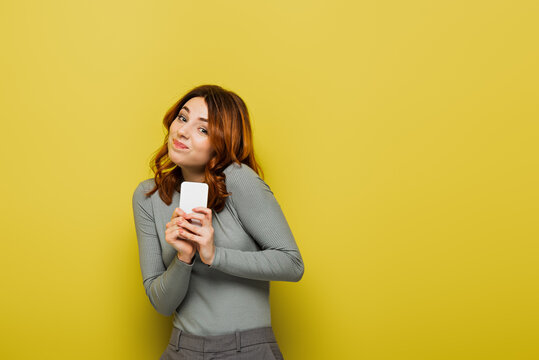 confused young woman with curly hair holding smartphone and looking at camera on yellow