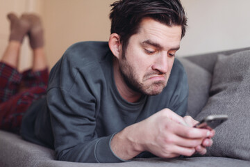 Fototapeta premium Man is lying on a couch with a smartphone, looks surprised and unhappy 