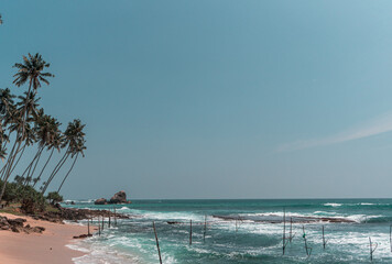 Tropikalna plaża z palmami, niebieski ocean z falami oraz kije rybackie.