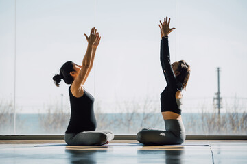 Two young women sitting the floor in lotus position, hands up and looking up, during yoga prenatal practice in a big sunny gym, fitness center. Sunny lighting indoors.