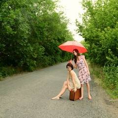 Young beautiful women with vintage suitcase trying to take a car hitch-hiking. Charming female travelers with retro bag and red umbrella, summer vacation