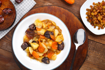 A dish of Jewish cuisine sweet tsimes with carrots and dates in a white plate on a round stand next to a spoon and carrots with dried fruits.