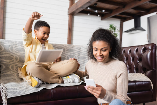 happy african american boy showing win gesture near mother with smartphone