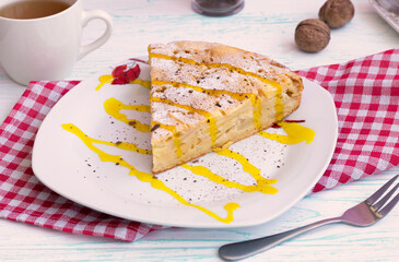 portion of vanilla pie on a plate. Decorated with powdered sugar, cinnamon and banana topping. kitchen napkin, with a fork and a cup of tea on a wooden table, nuts in background. wood background