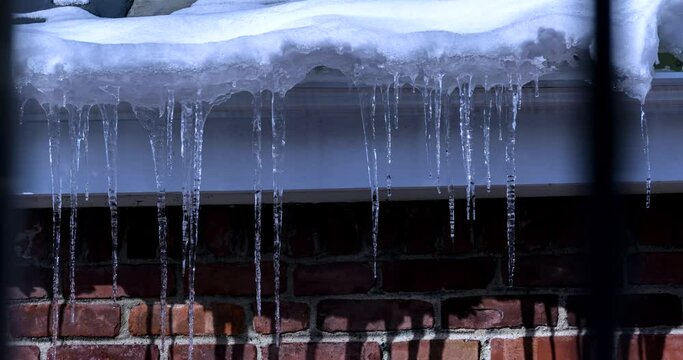 A 6 hour long time-lapse of winter icicles and white snow handing from a rooftop in front of a red brick wall and melting.